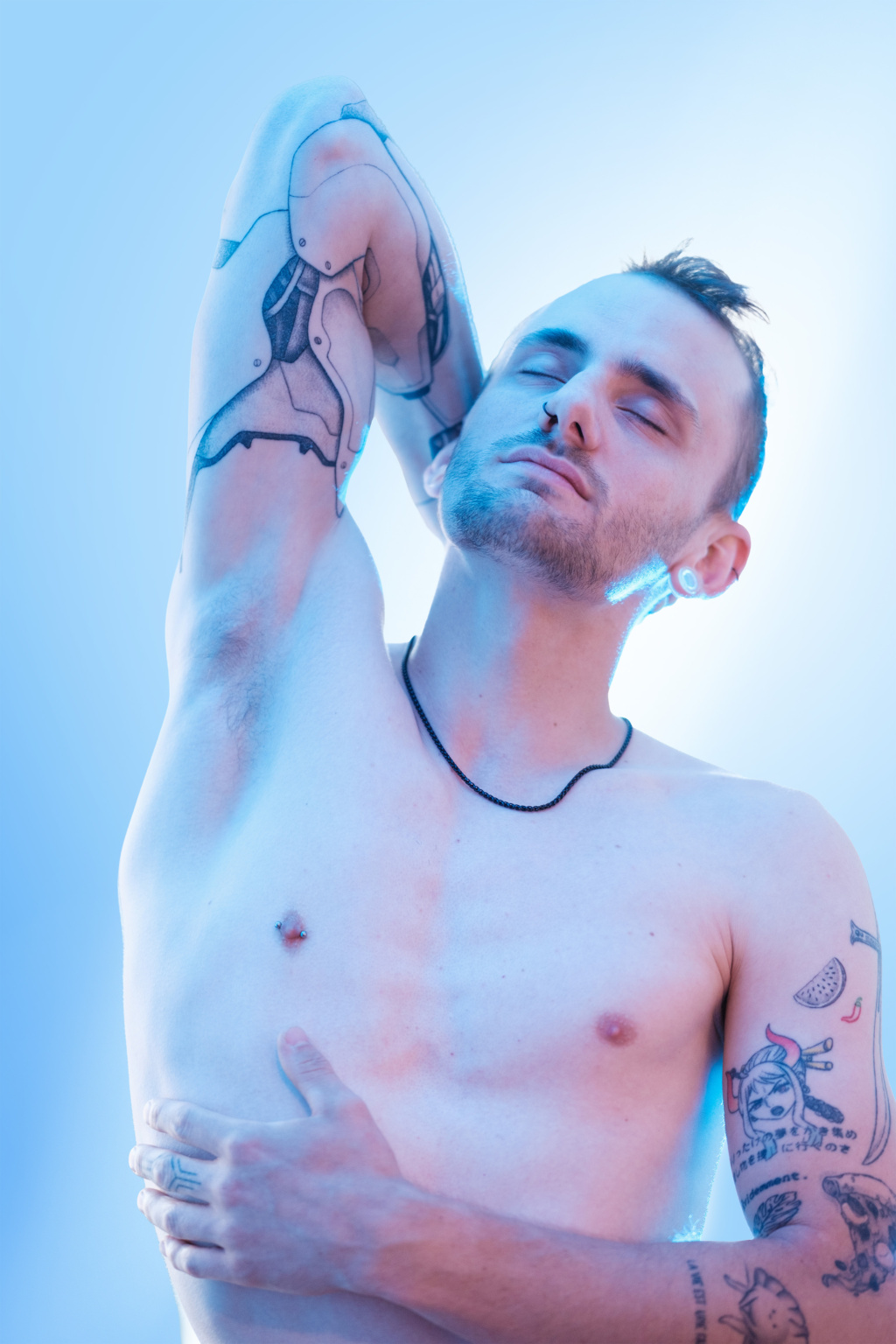 Portrait artistique avec lumière bleutée d'un homme torse nu, bras levé révélant un tatouage de bras robotique façon manga, collier fin et piercings, ambiance studio épurée