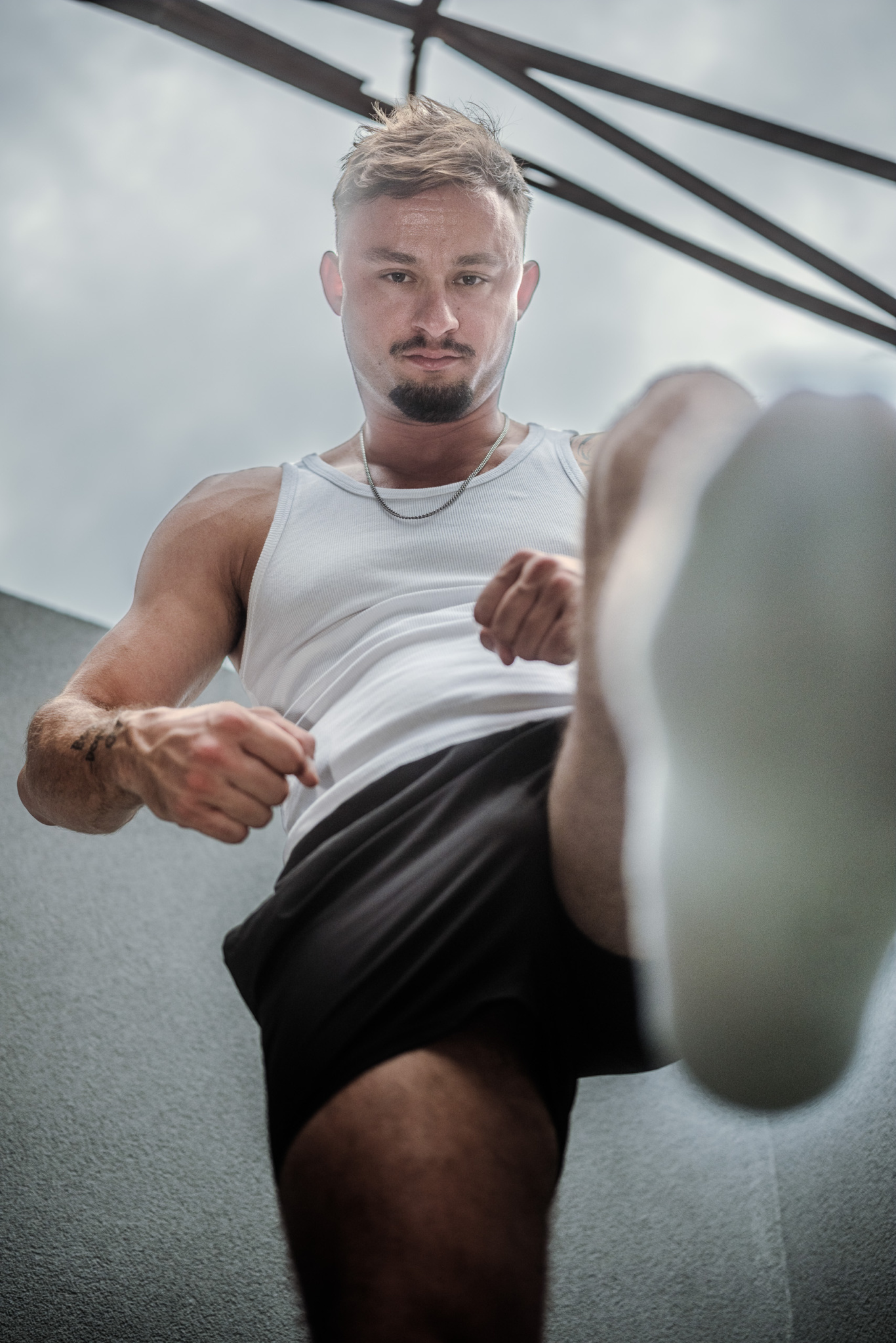 Photo de KevinHungXL devant un mur extérieur, en débardeur et short noir, posture dominante avec chaussettes