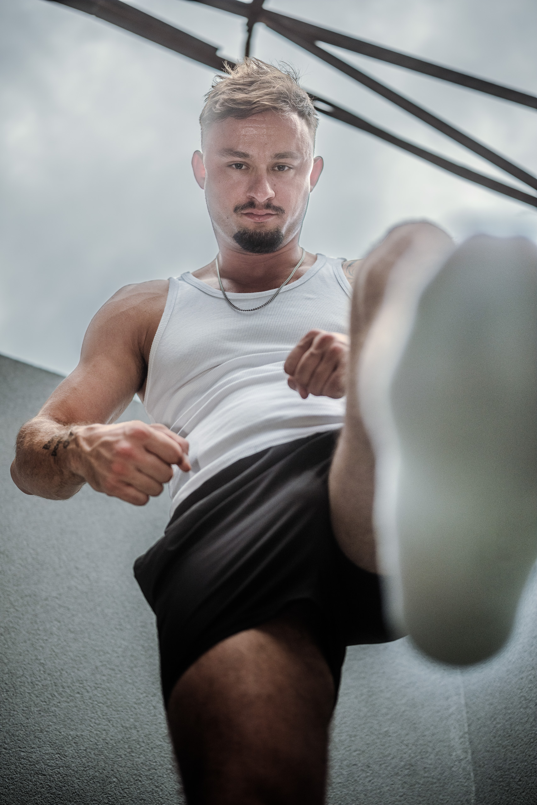 Photo de KevinHungXL devant un mur extérieur, en débardeur et short noir, posture dominante avec chaussettes