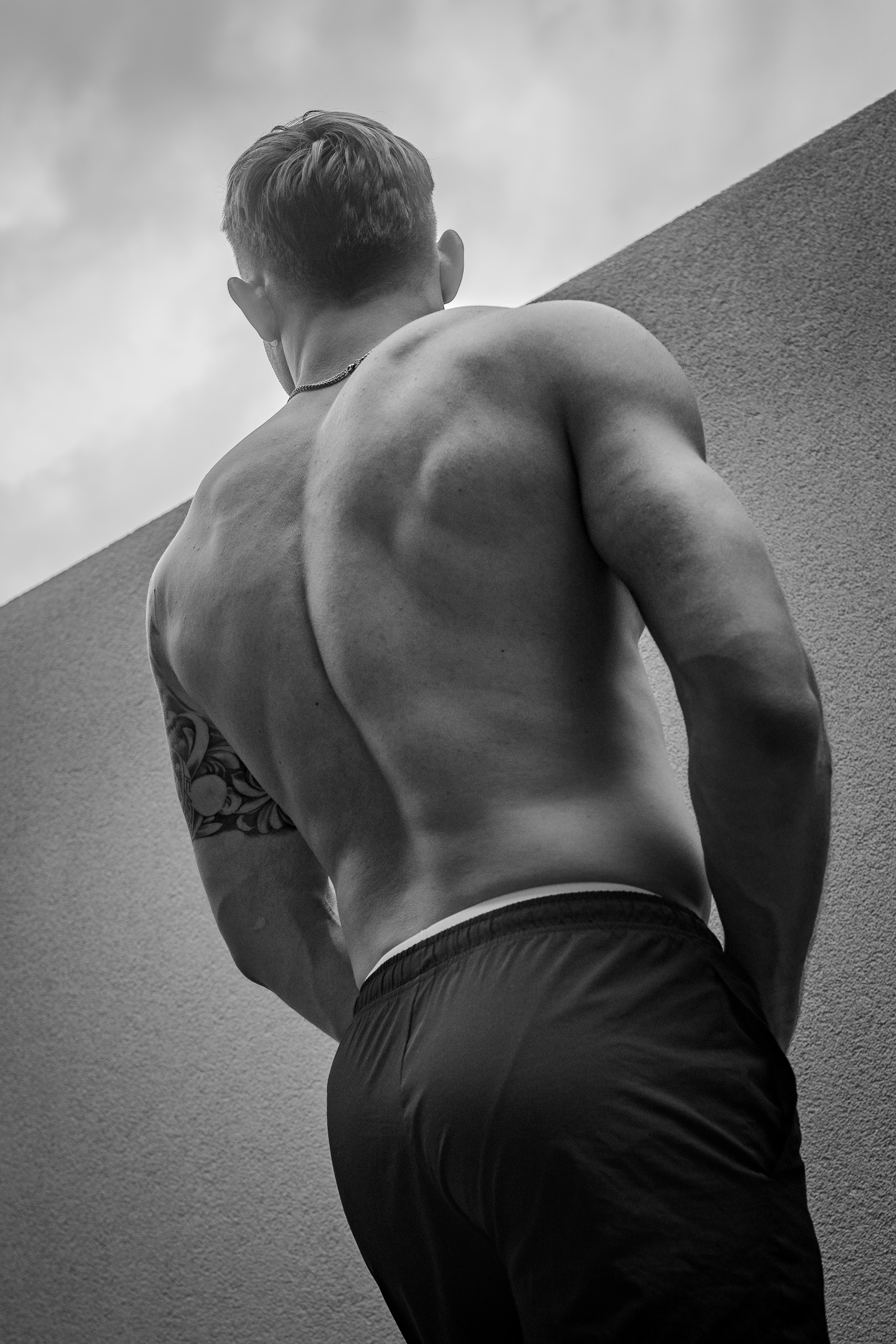 Photo de dos de KevinHungXL en short noir, devant un muret gris, dos musclé