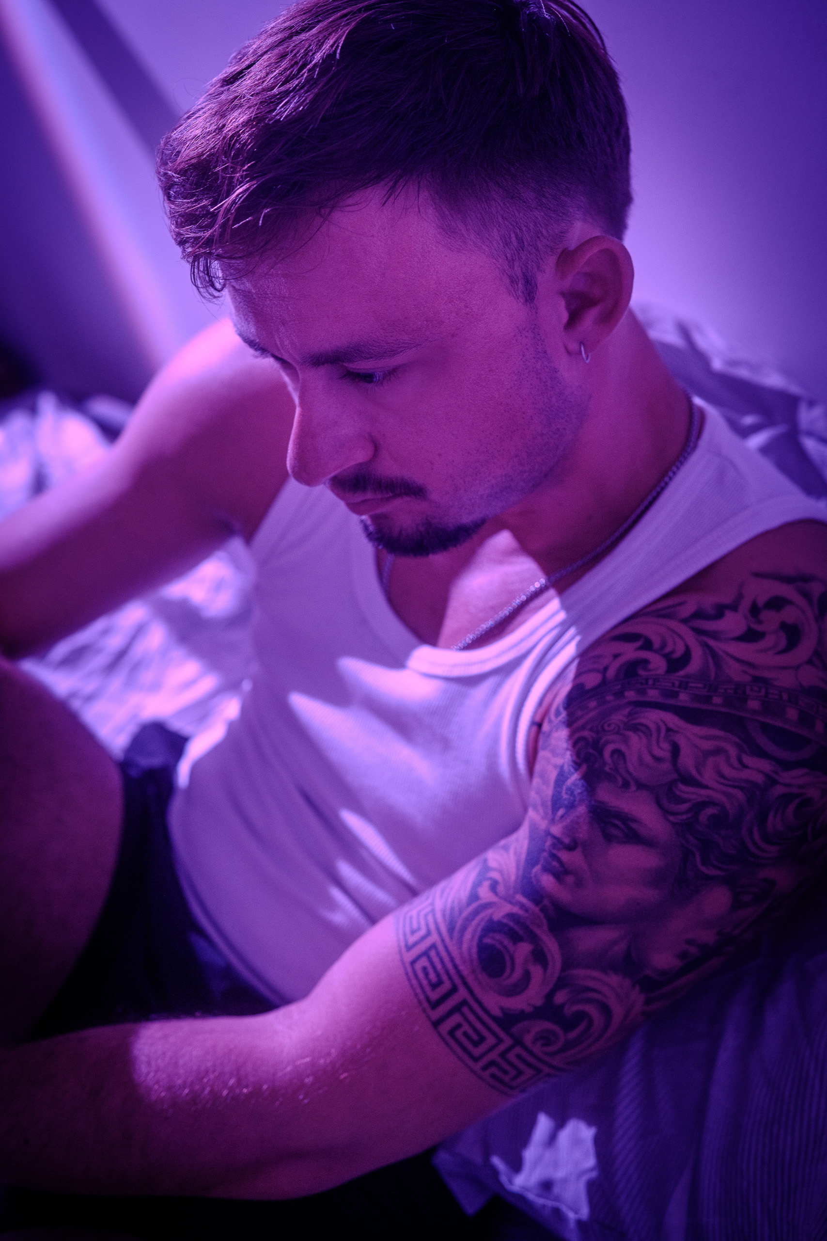 Portrait de KevinHungXL, éclairage rose et violet, assis sur un lit en short de sport et débardeur blanc, vue sur son tatouage grec