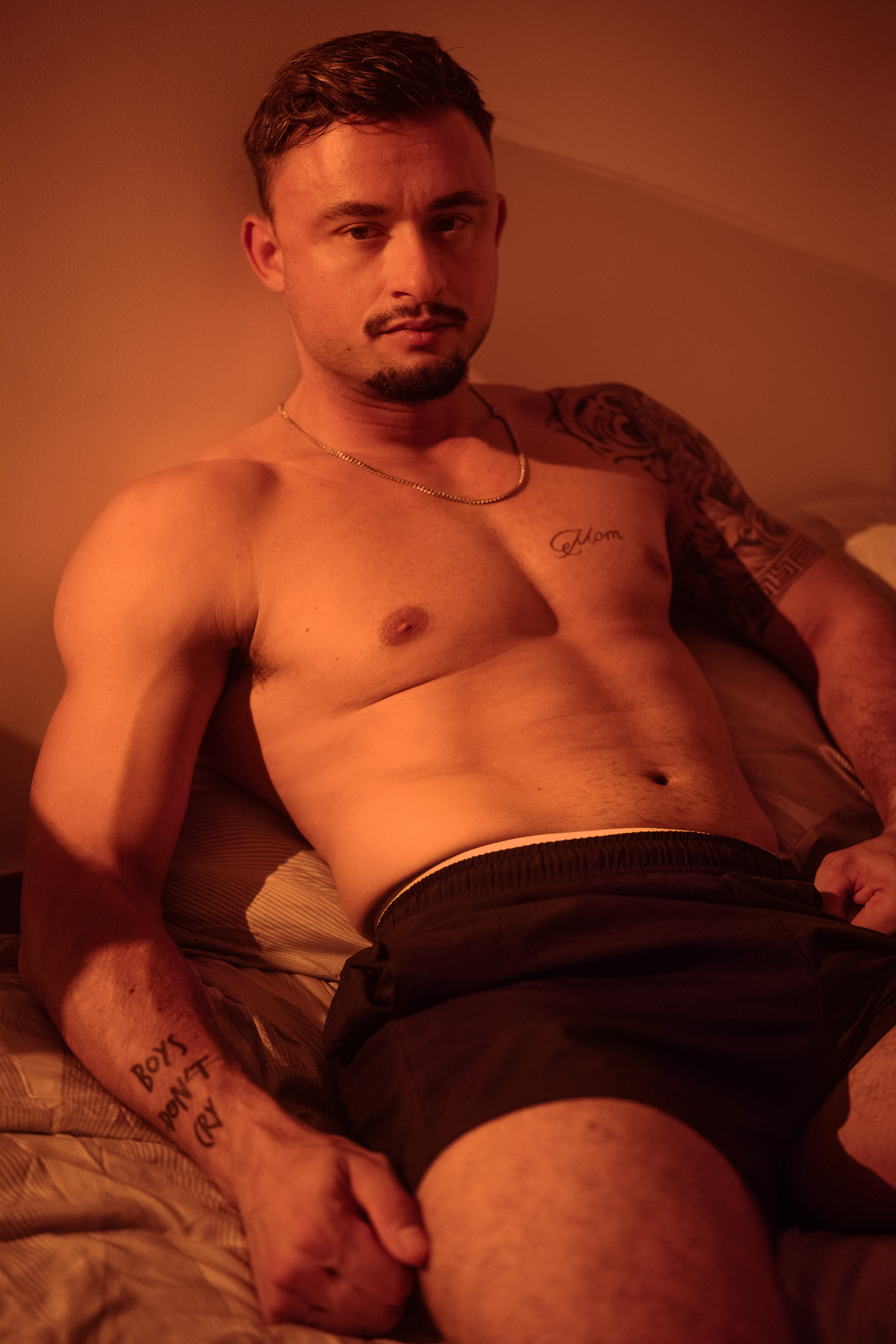Portrait de KevinHungXL en short de sport et torse musclé, assis sur un lit, avec éclairage en tons chauds