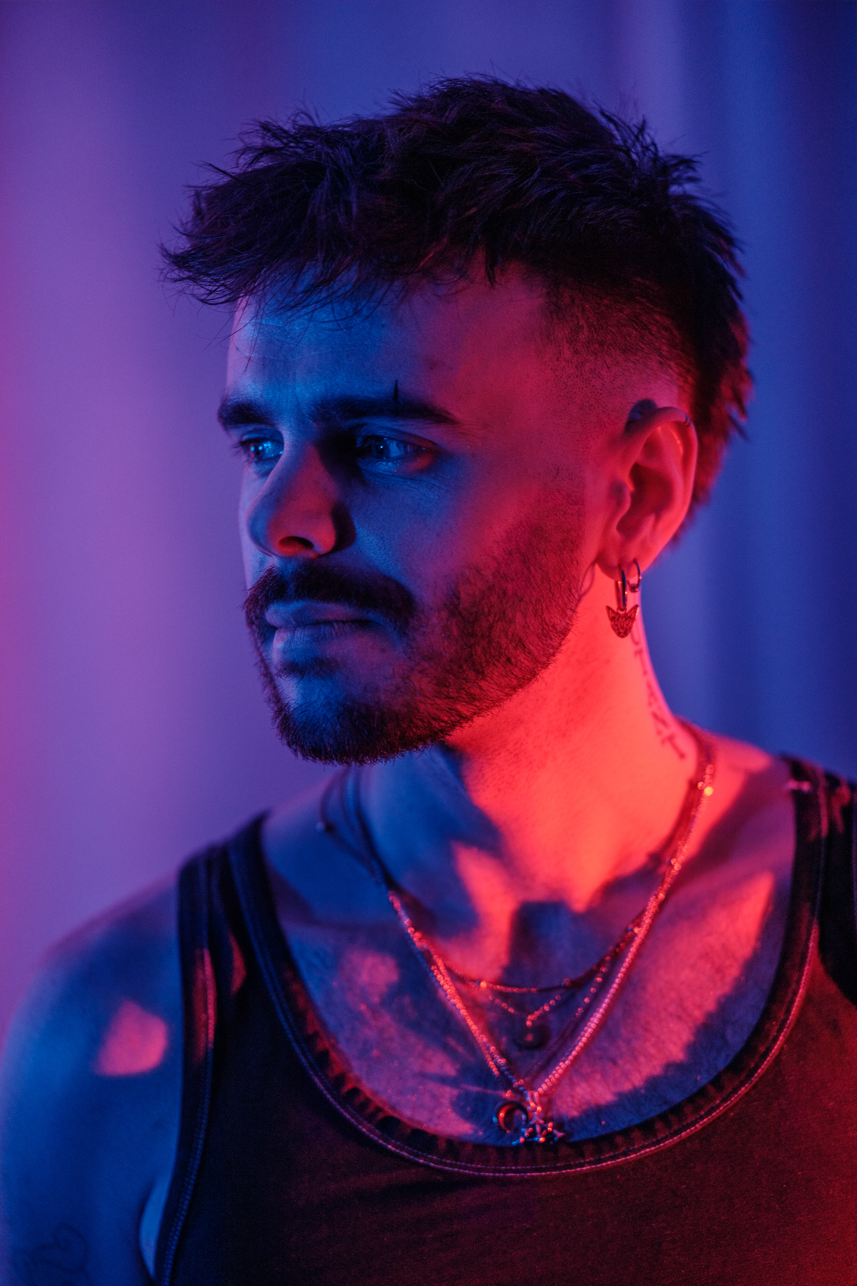 Portrait de profil, bijoux et tatouage au cou, éclairage violet et rose.