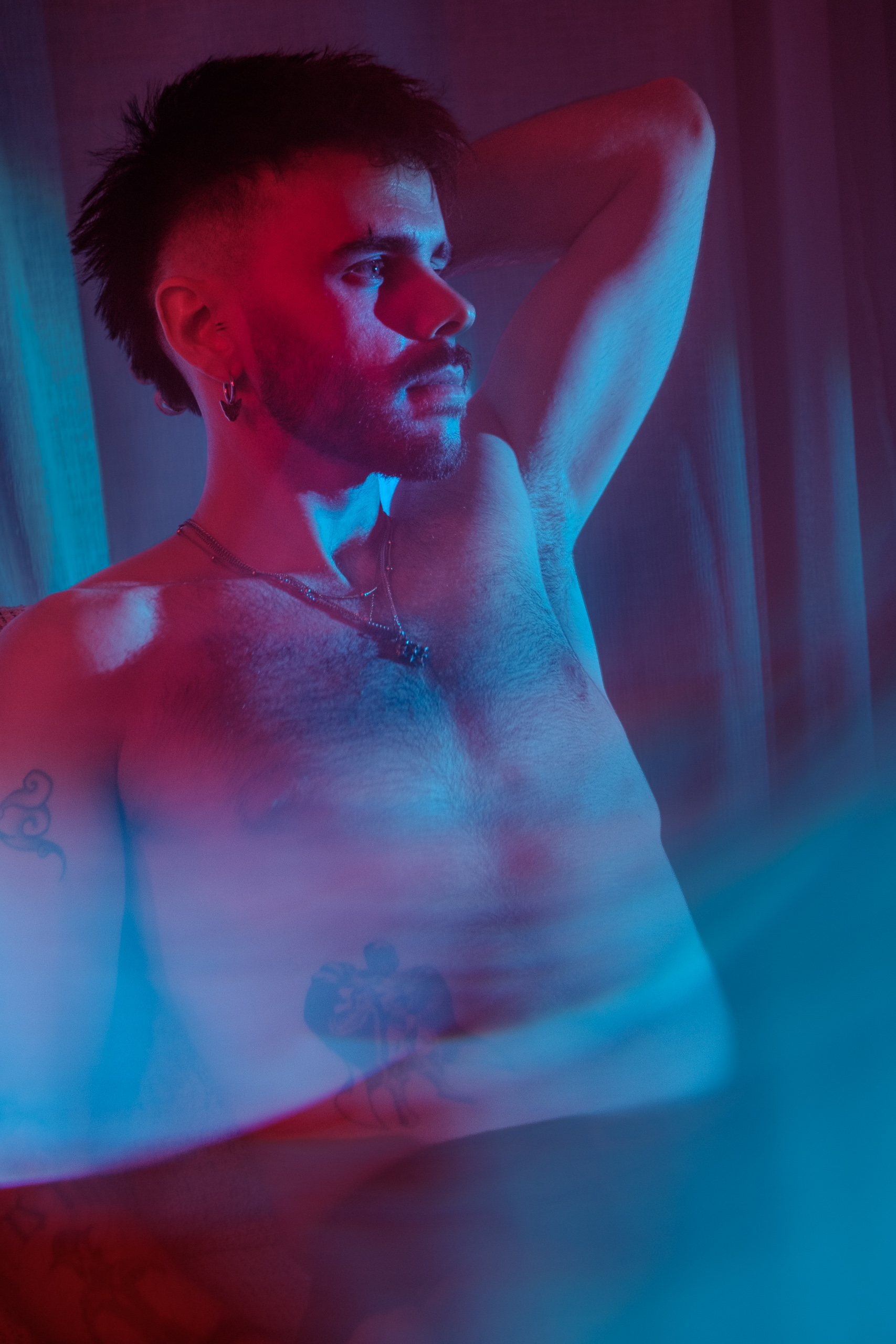 Portrait torse nu, bras levé, tatouages, éclairage bleu et rose.