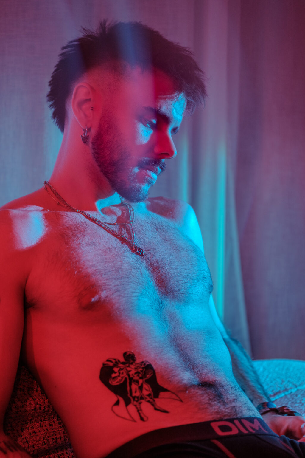 Portrait torse nu assis, tatouage abdominal, éclairage rouge et bleu.
