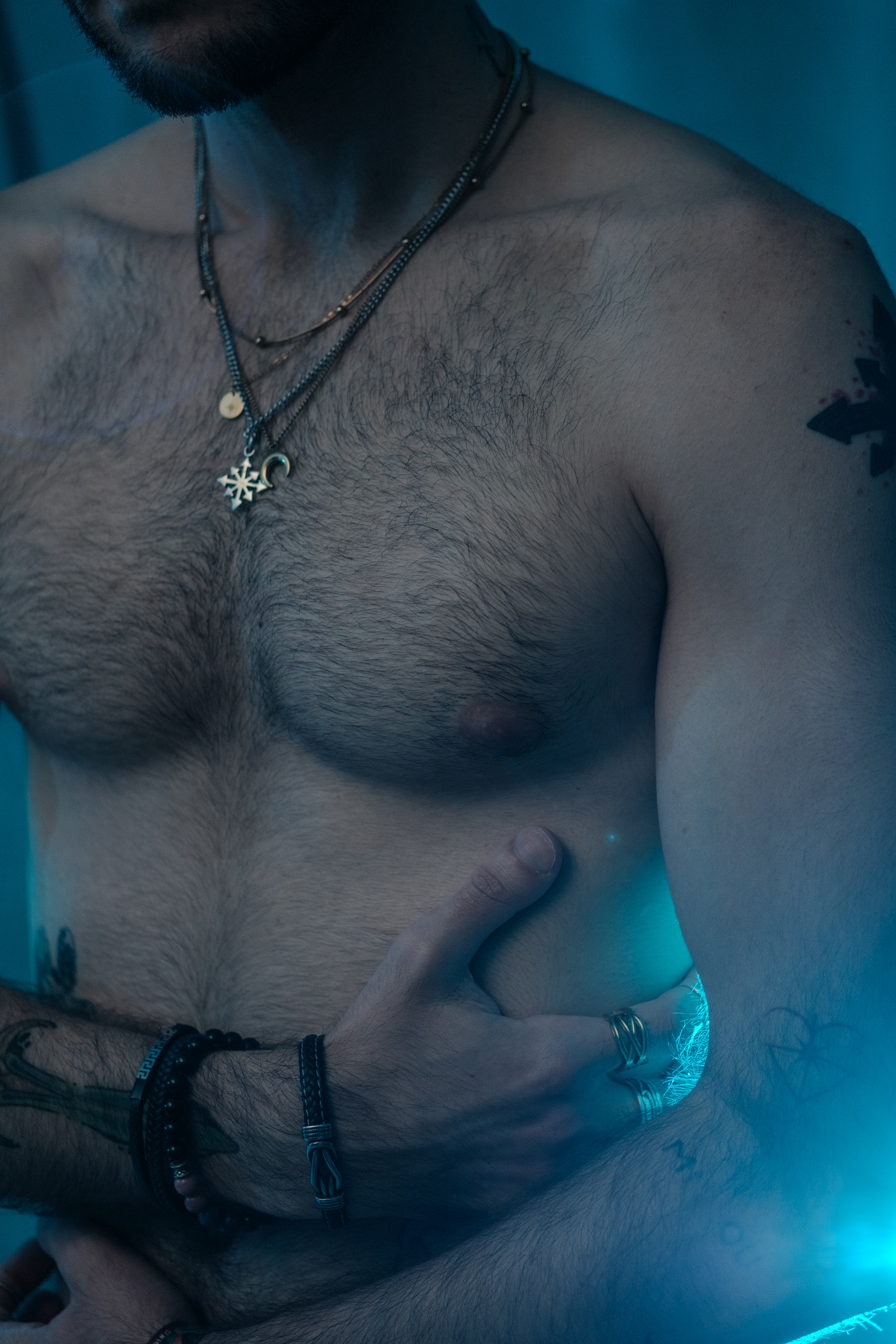 Gros plan sur le torse, colliers et bagues, éclairage bleu tamisé.