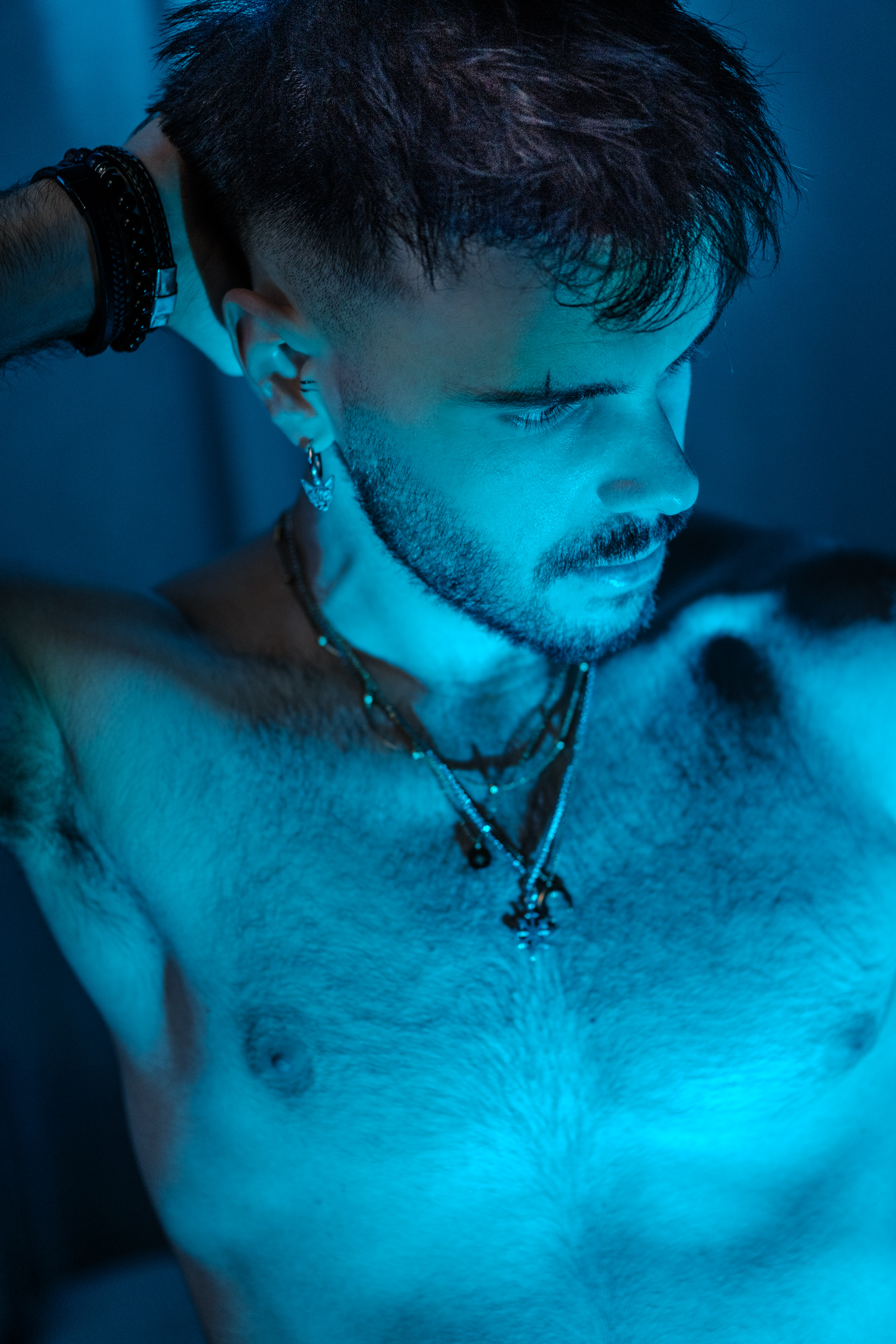 Portrait torse nu, colliers, éclairage bleu, regard baissé.