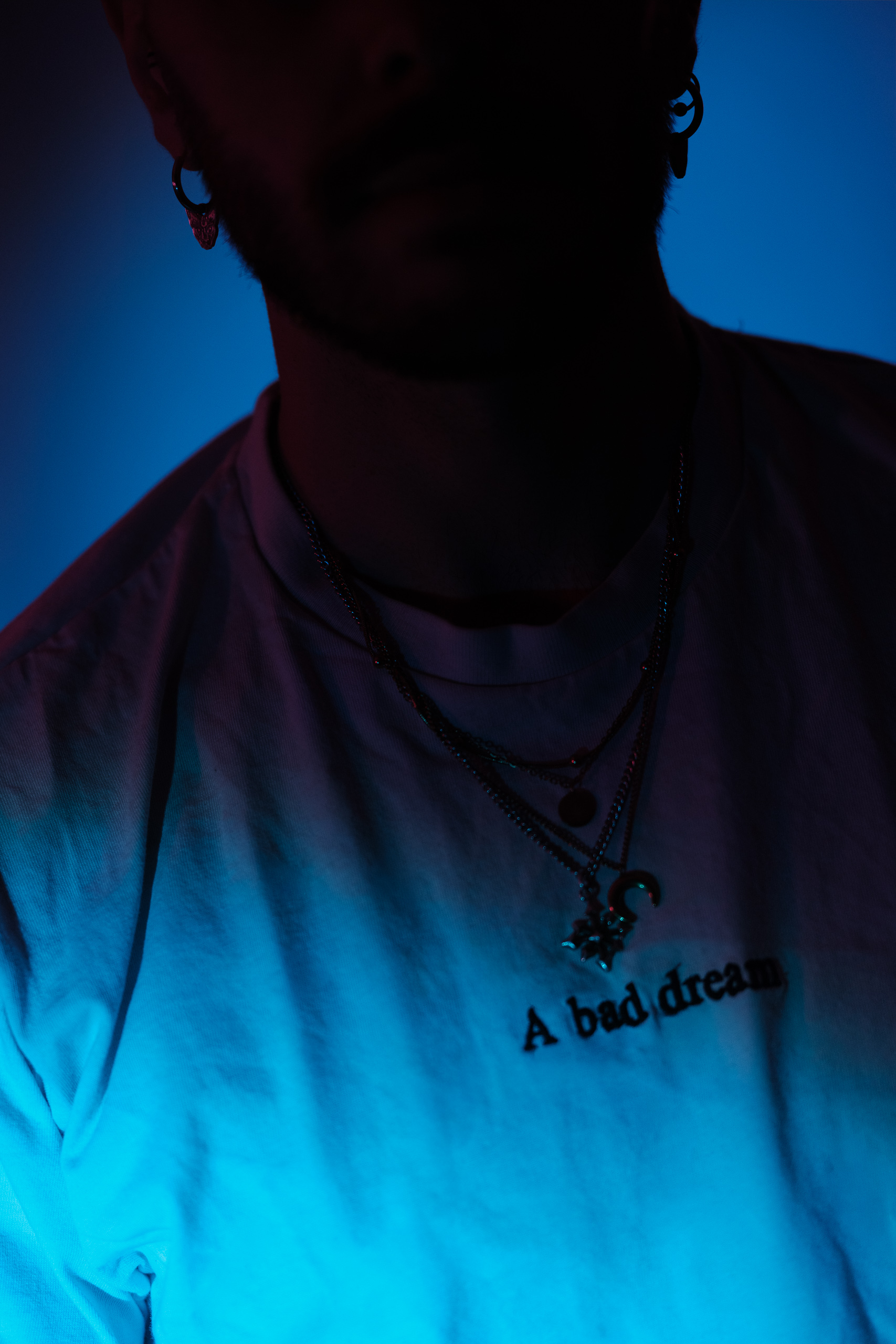 Portrait rapproché, t-shirt et colliers, éclairage bleu tamisé.