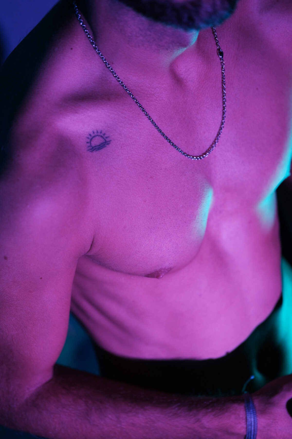 Loukas More, gros plan épaule et buste avec tatouage soleil, lumière violette, portrait artistique masculin.