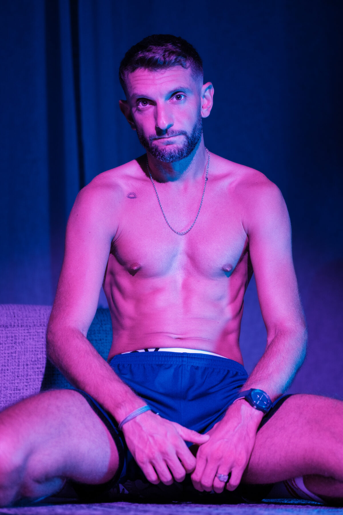 Loukas More assis en short marine, bague et montre, ambiance rose et bleu, portrait lifestyle.