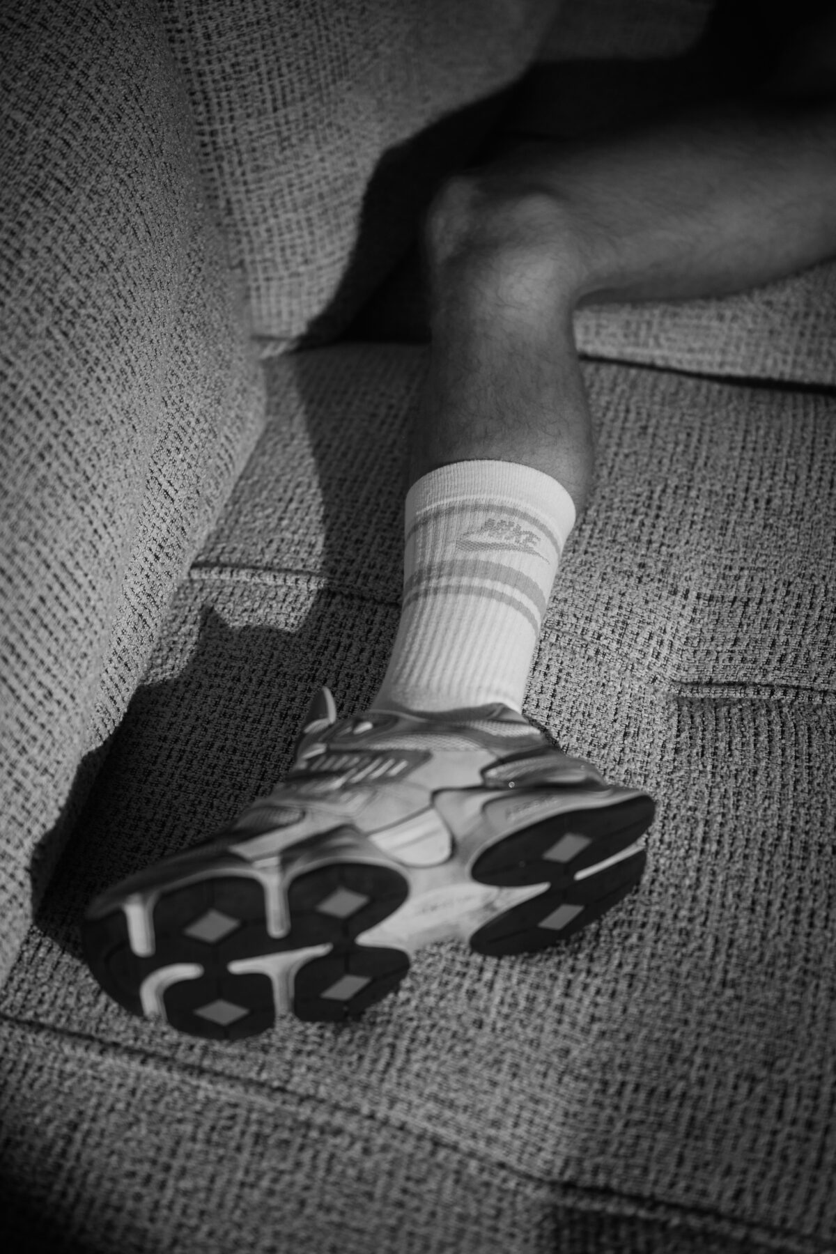 Détail chaussette et sneaker sur canapé, jambe de Loukas More, photographie noir et blanc lifestyle.