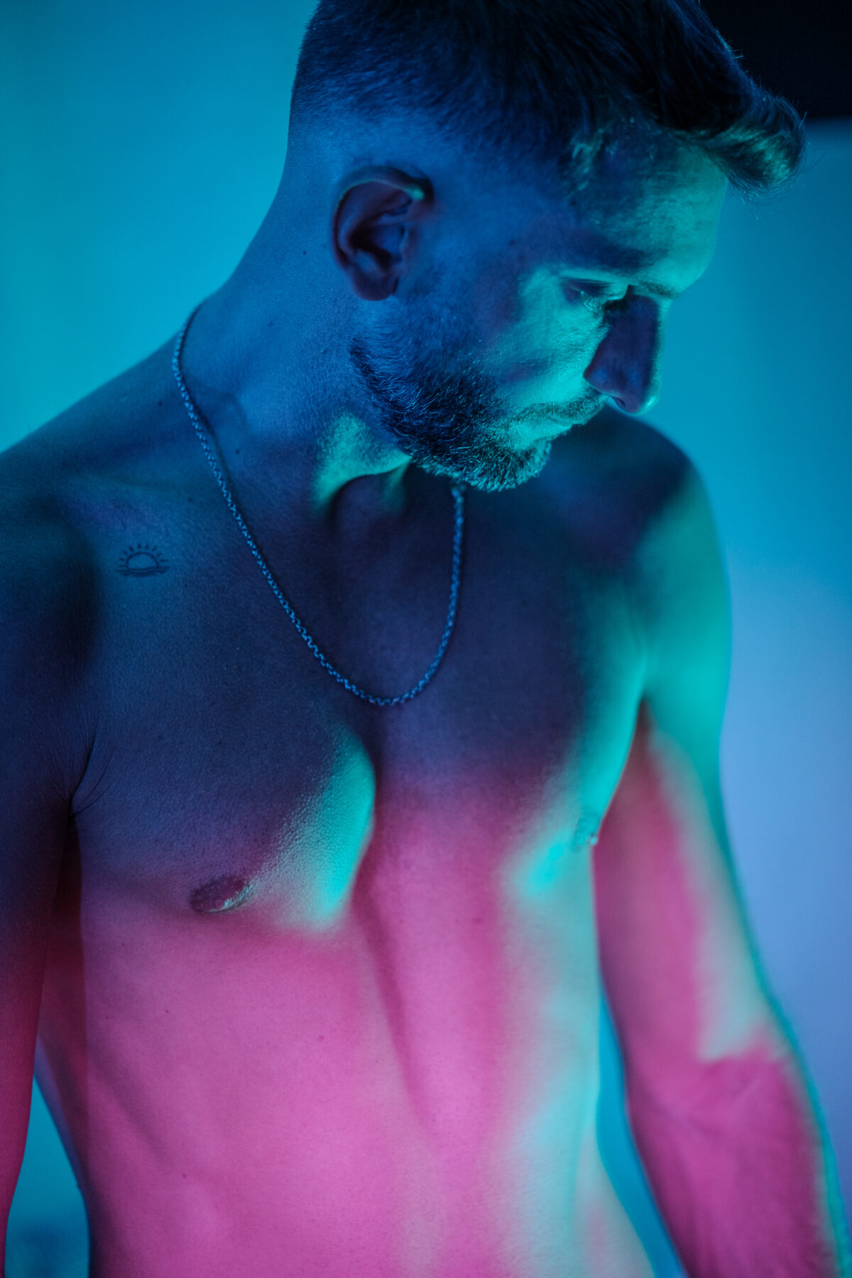 Portrait torse nu de Loukas More, chaîne en argent, éclairage bicolore bleu et rose, photographie de mode homme.