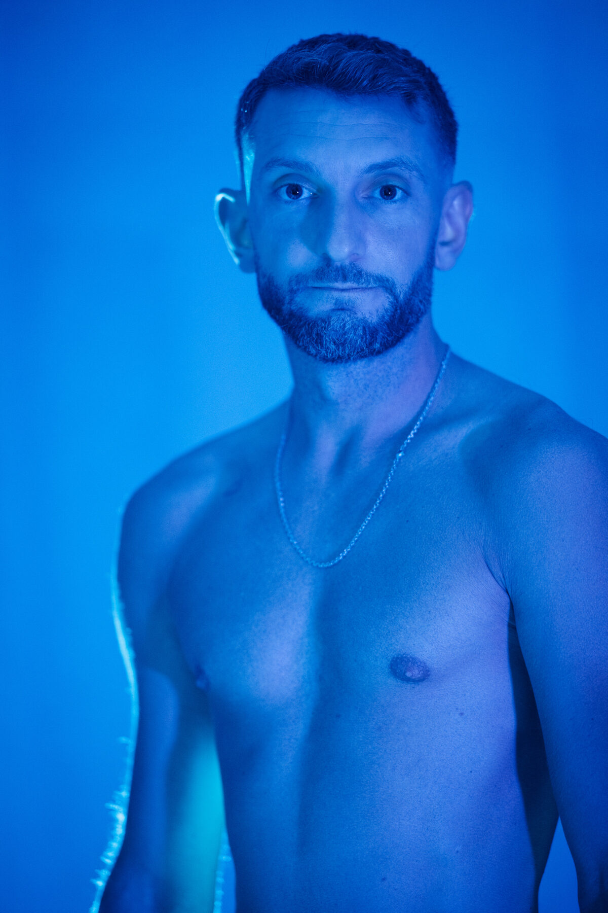 Portrait de Loukas More, visage et torse, fond bleu profond, regard caméra, photographie masculine épurée.