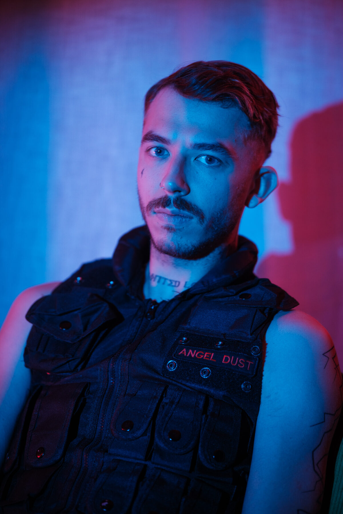Portrait bicolore bleu et rouge d'Angel Dust, gilet tactique floqué à son nom, tatouages au cou et au bras, regard direct.