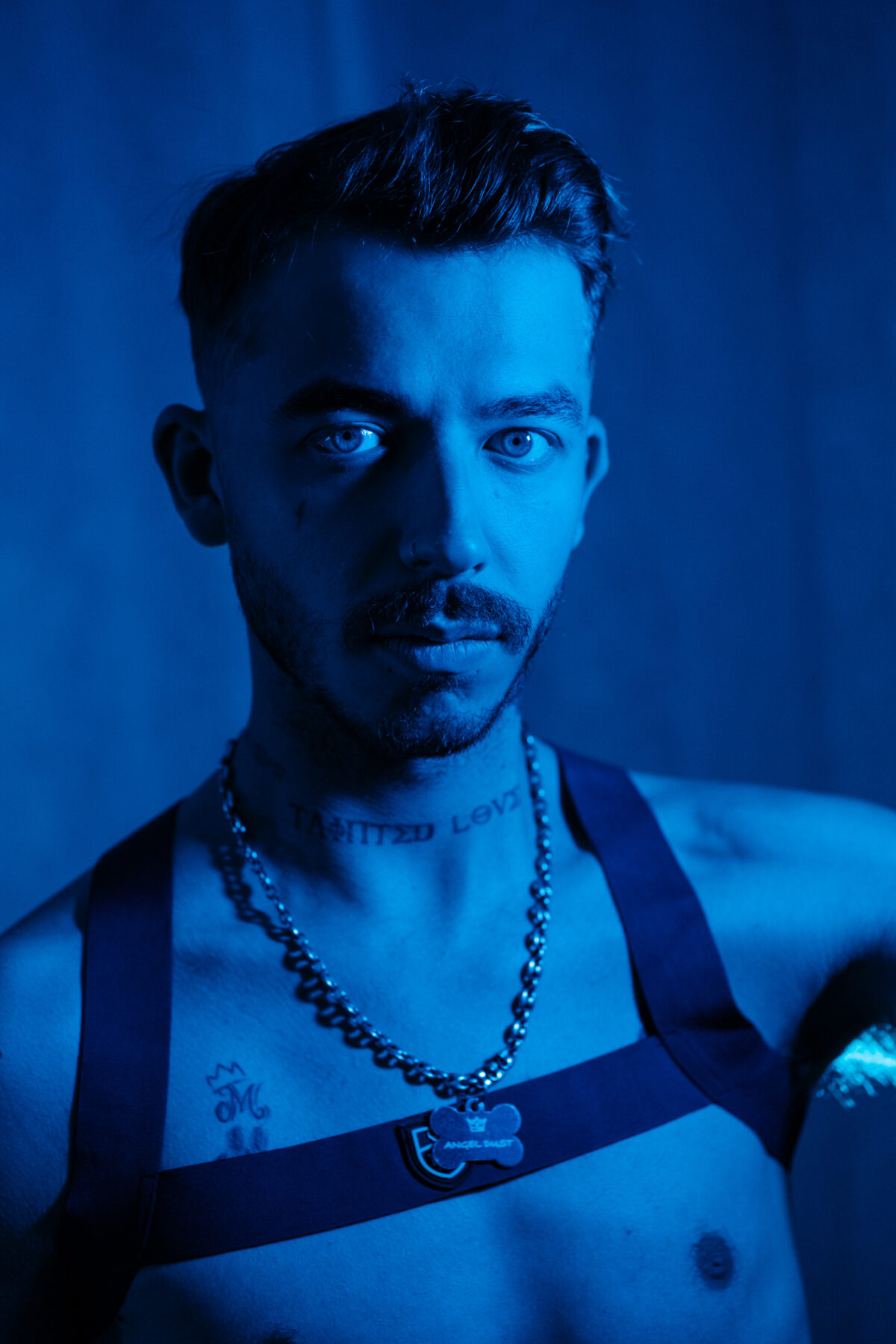 Portrait bleu d'Angel Dust, harnais à médaille "Angel Dust", collier chaîne, tatouage "Tainted Love" au cou, regard caméra.
