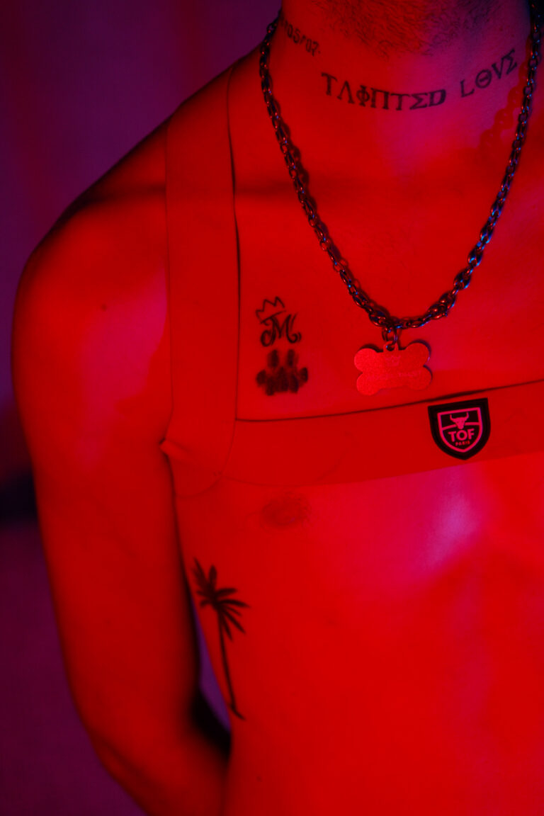Portrait bicolore rouge et bleu d'Angel Dust, débardeur à logo, collier chaîne, tatouage "Tainted Love" au cou.