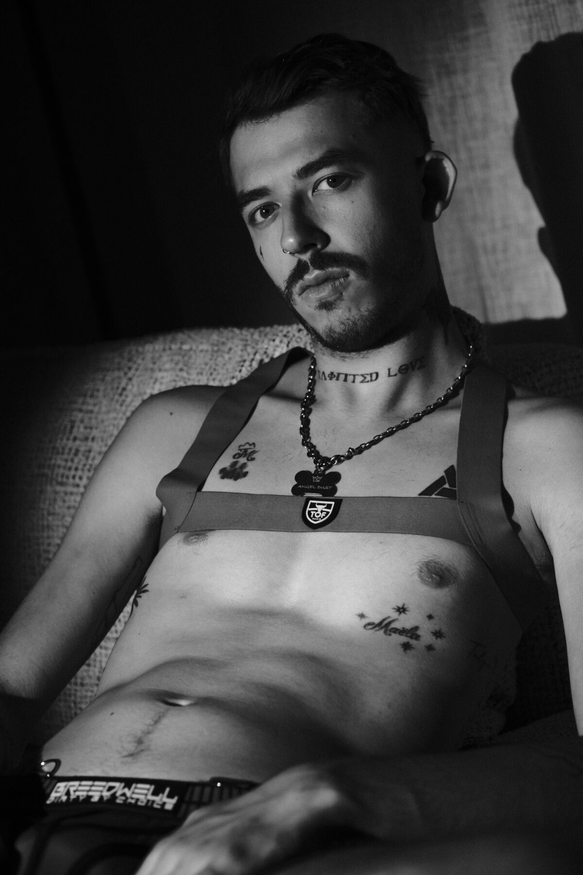 Portrait noir et blanc d'Angel Dust assis, harnais à médaille "Angel Dust" et écusson "TOF Paris", tatouages, regard caméra.