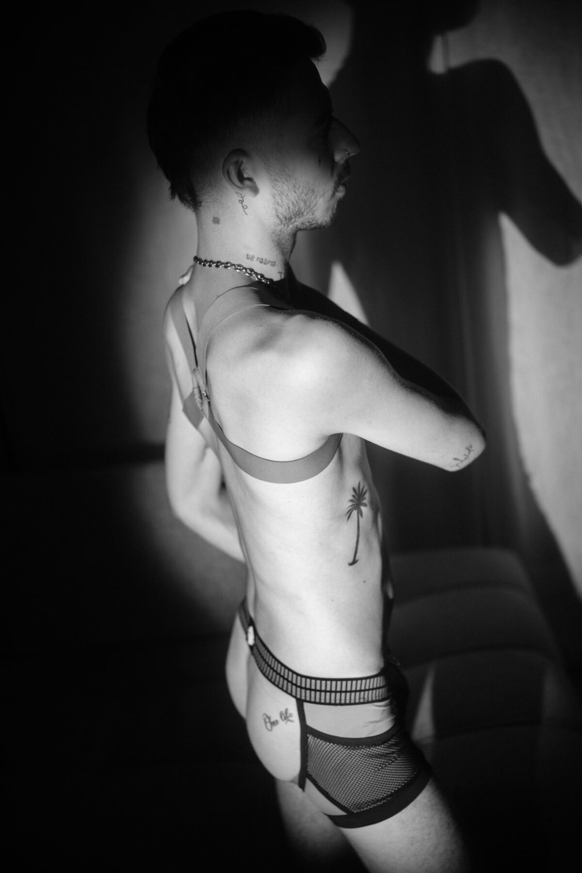 Profil noir et blanc d'Angel Dust, harnais et jockstrap, tatouages (palmier, texte) sur les côtes et le bras.