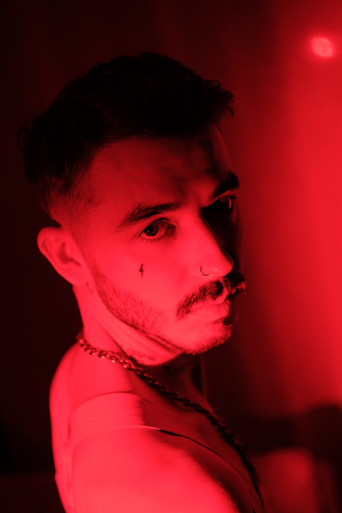 Portrait rouge en profil d'Angel Dust, chaîne au cou, piercing au nez, regard dirigé hors champ.