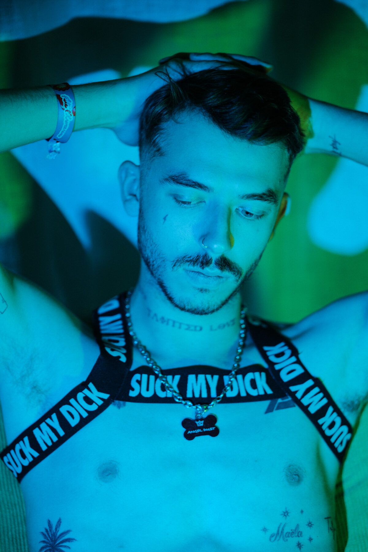 Angel Dust bras levés derrière la tête, harnais "Suck my dick", bracelet, tatouages (palmier, étoiles, prénom).