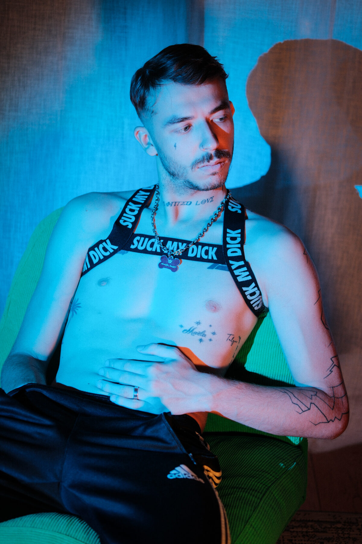 Angel Dust assis, main sur le ventre, harnais "Suck my dick", bague, tatouages, short Adidas en arrière-plan.