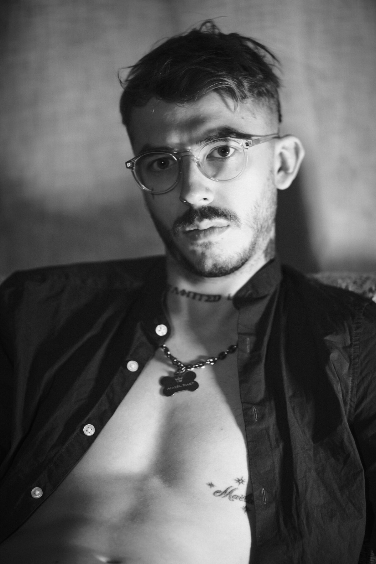 Portrait noir et blanc d'Angel Dust avec lunettes rondes, chemise ouverte, médaille "Angel Dust", tatouage "Tainted Love" au cou.