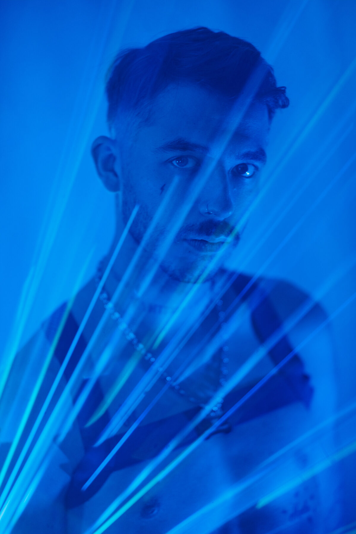 Portrait bleu d'Angel Dust voilé par des fils lumineux en premier plan, collier à chaîne, regard caméra.