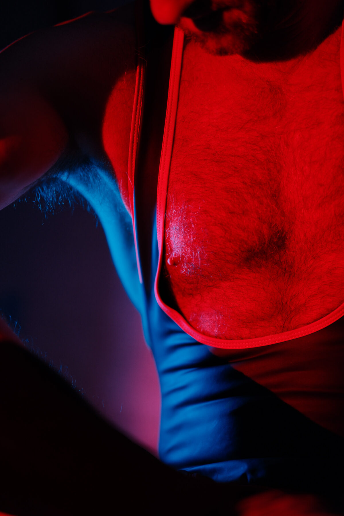 Gros plan sur un torse velu portant un harnais, contraste de lumière rouge et bleue