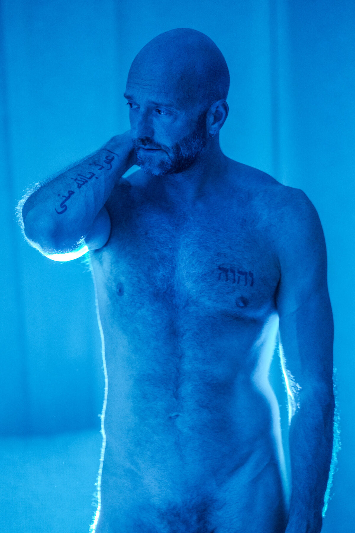 Nu artistique en pied de Benoit sous une lumière bleue dramatique, tatouages hébreu et arabe visibles