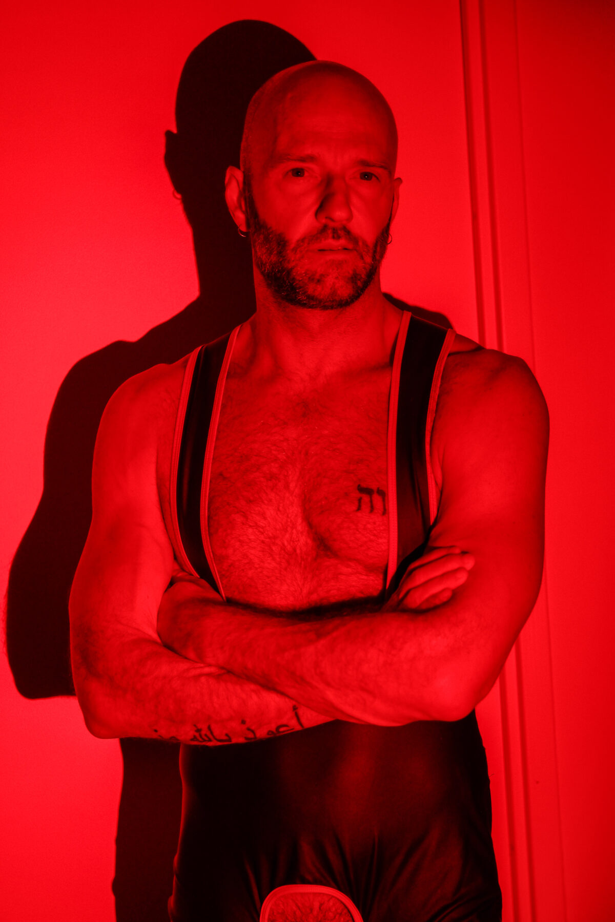 Portrait de Benoit debout, bras croisés, harnais noir sous lumière rouge, tatouages hébreu et arabe visibles