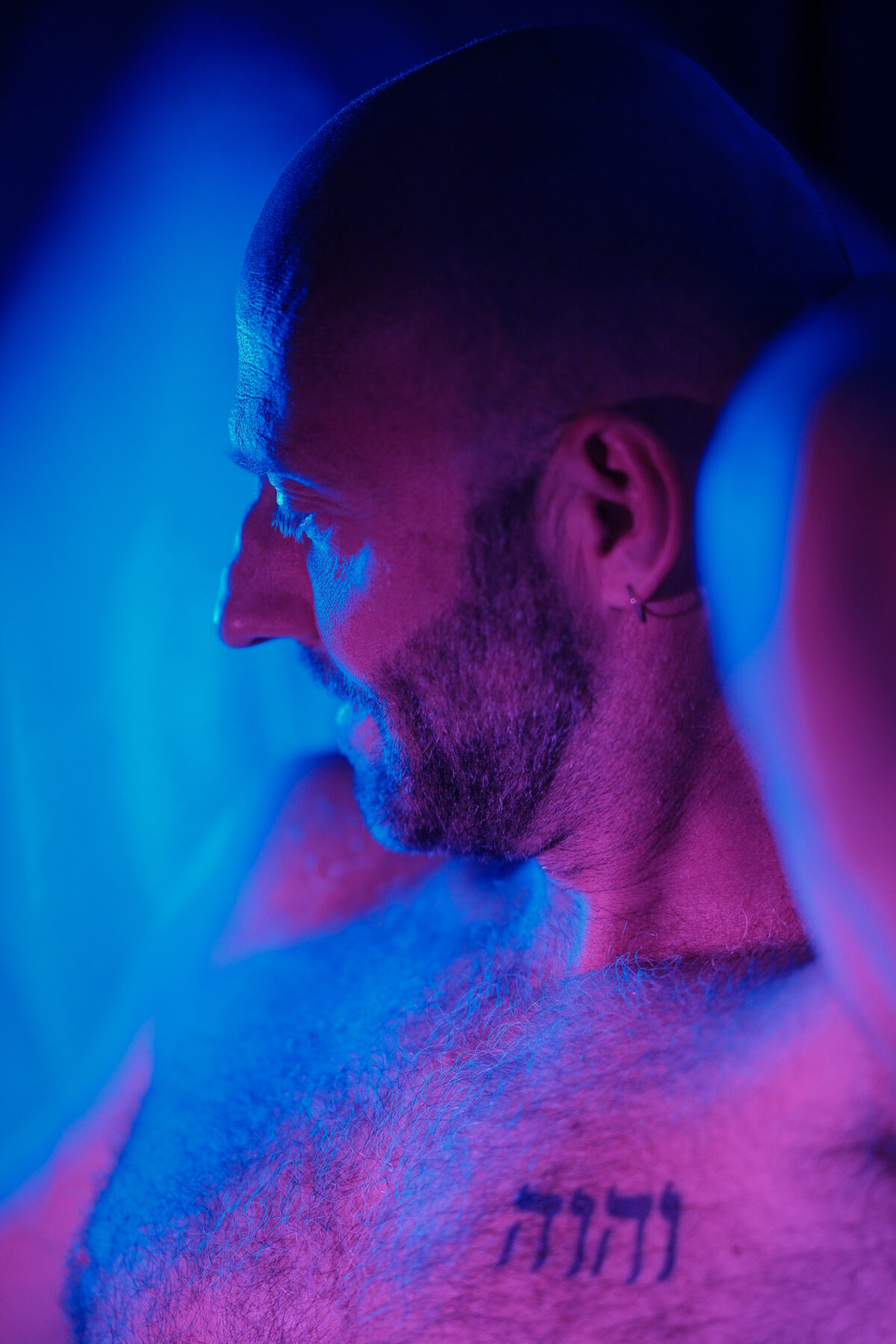 Portrait de profil de Benoit, crâne rasé et barbe taillée, tatouage hébreu sur le torse, lumière rose et bleue
