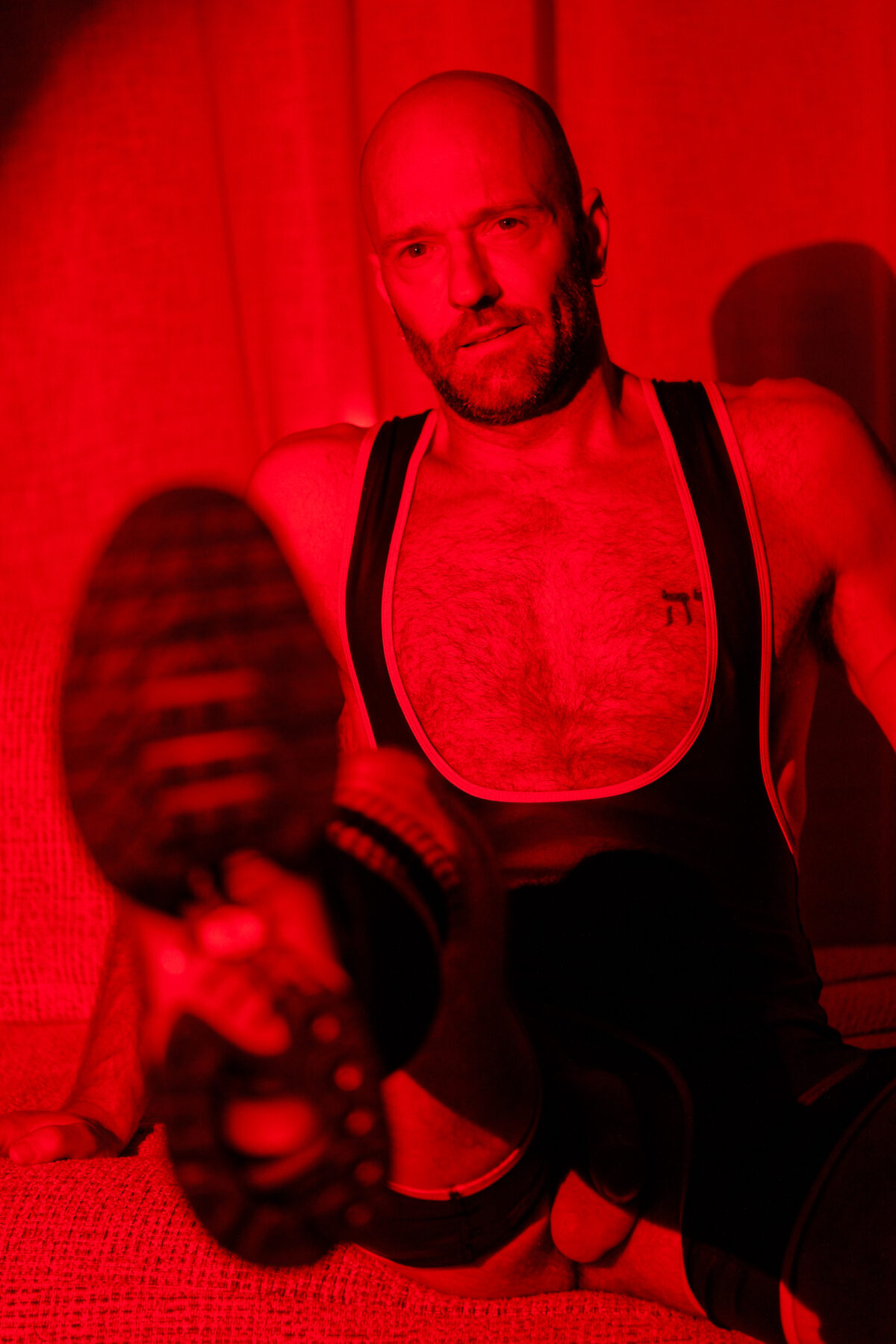 Portrait de Benoit sous lumière rouge, assis en harnais noir, jambe tendue vers l'objectif, ambiance ludique