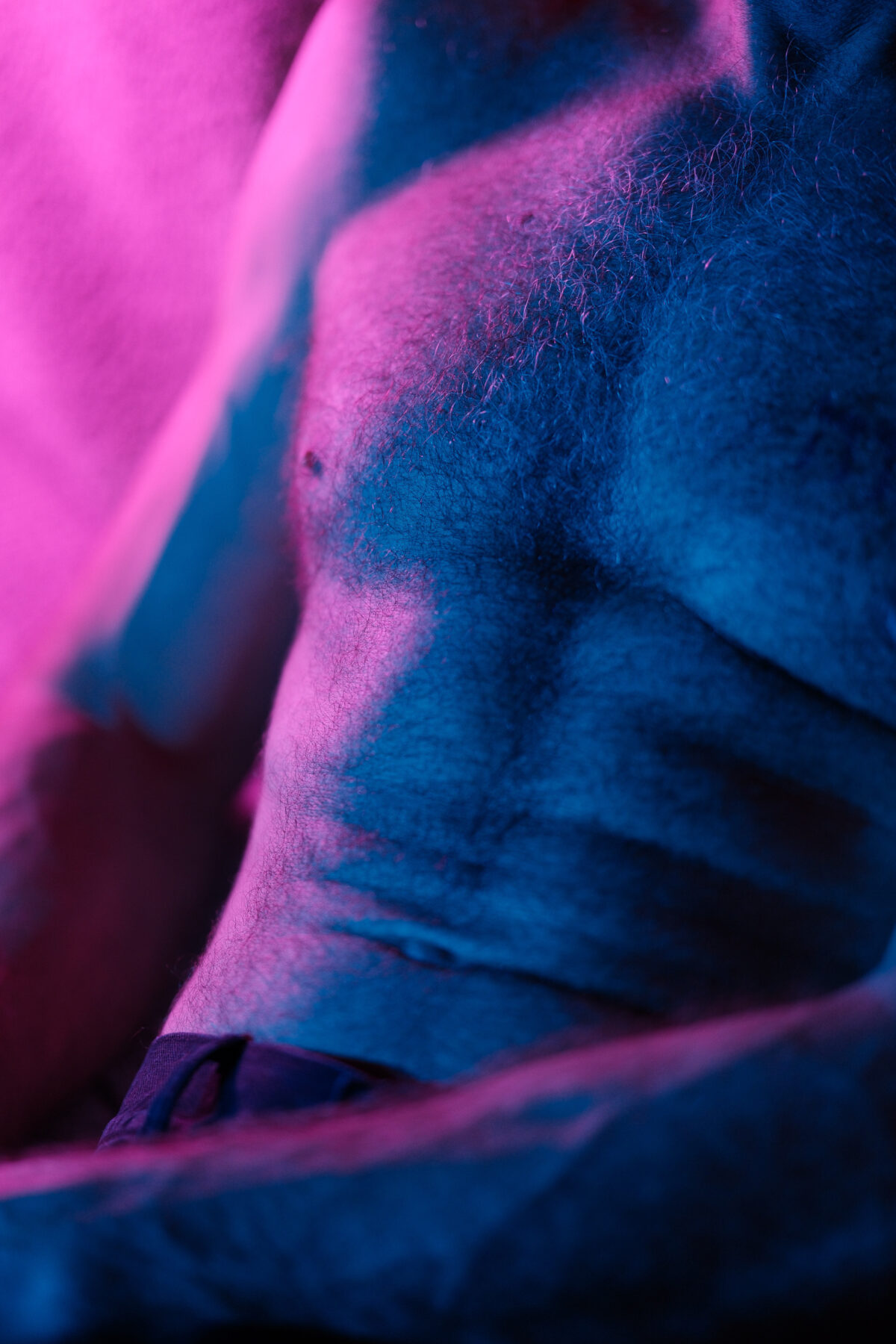 Gros plan abstrait sur la peau et la pilosité d'un torse masculin, éclairage colorimétrique rose et bleu, photographie artistique