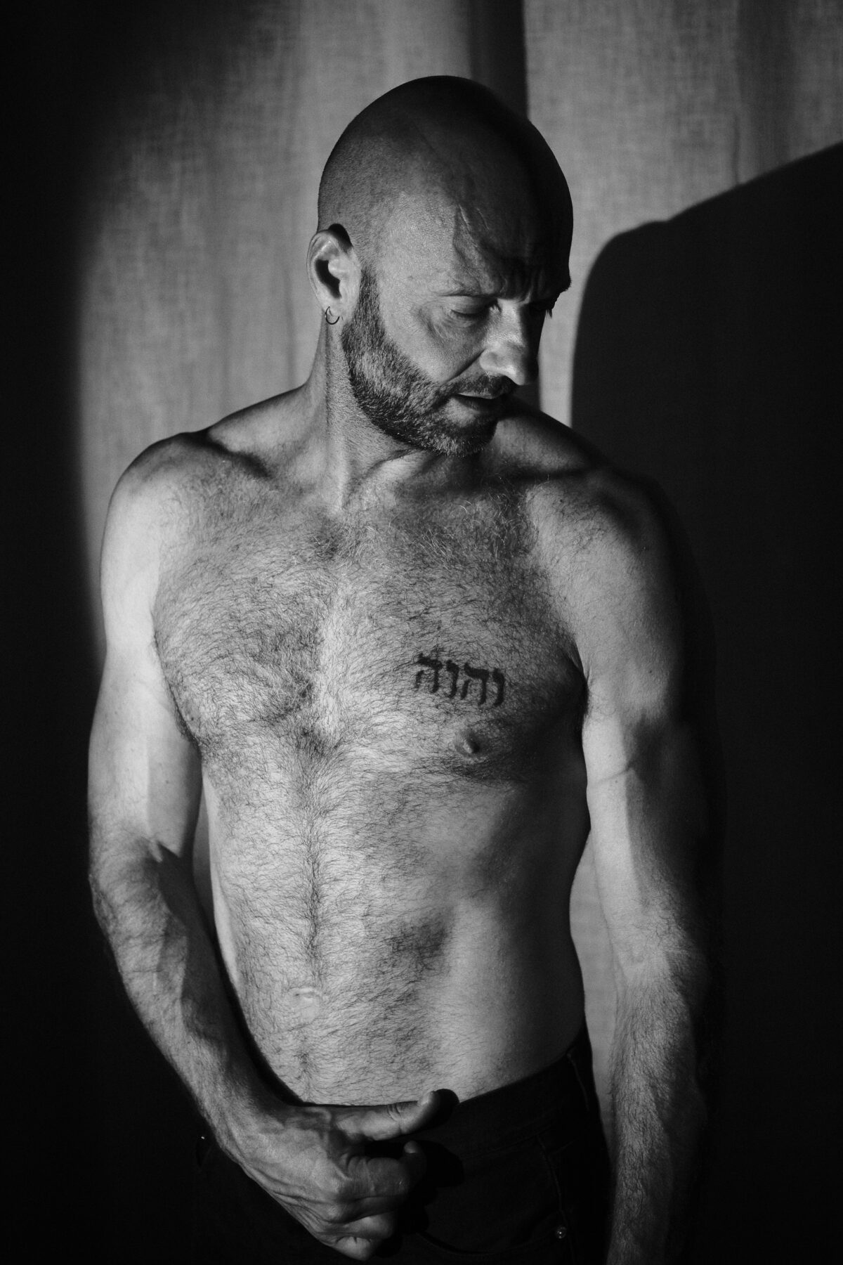Portrait noir et blanc de Benoit, torse nu et tatouage hébreu, lumière naturelle et rideau en arrière-plan