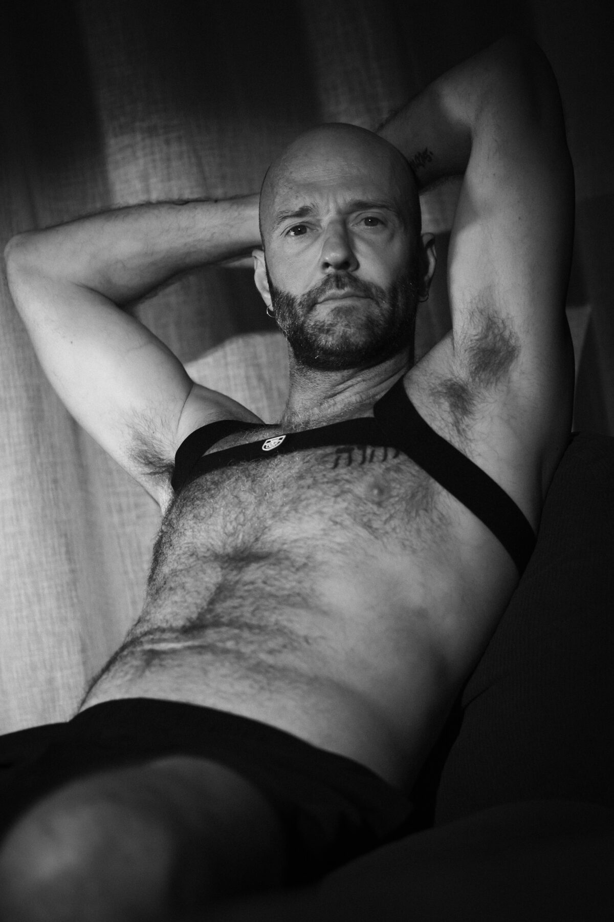 Portrait noir et blanc de Benoit, bras levés derrière la tête, harnais en cuir sur le torse, regard direct