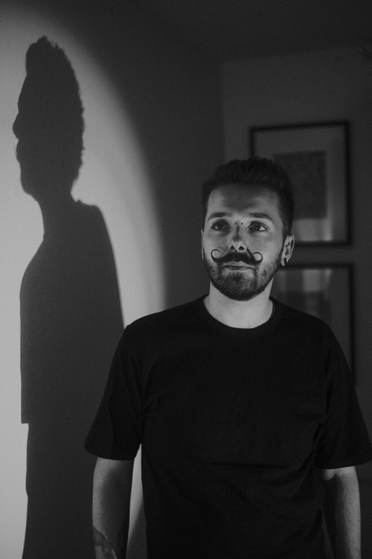 Portrait noir et blanc de François avec son ombre projetée sur le mur, cadres photo en arrière-plan, tee-shirt noir