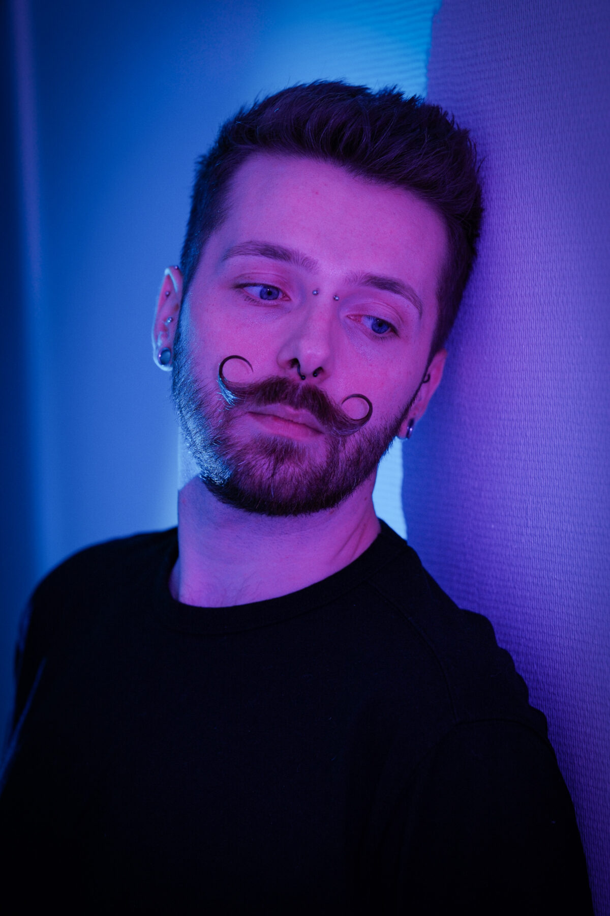 Portrait de François sous une lumière bleu-violet, regard baissé, piercings au visage, haut sombre