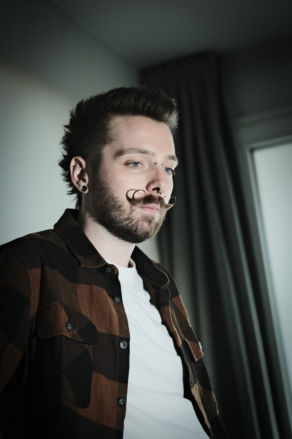 Portrait de François en chemise à carreaux ouverte sur un tee-shirt blanc, regard vers l'objectif, rideau et fenêtre en arrière-plan