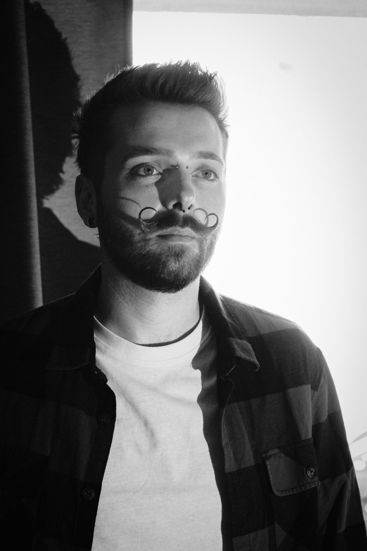 Portrait noir et blanc de François en chemise à carreaux ouverte, tee-shirt blanc, regard de côté