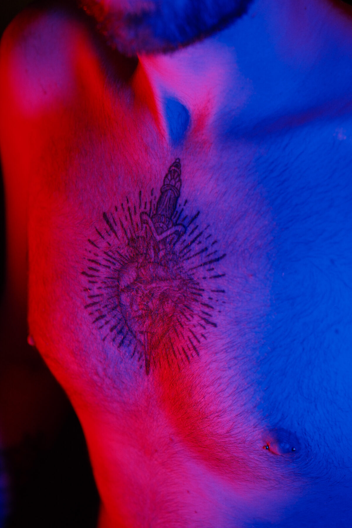 Gros plan sous lumière rouge et bleue sur le tatouage de François, cœur sacré encré sur le torse