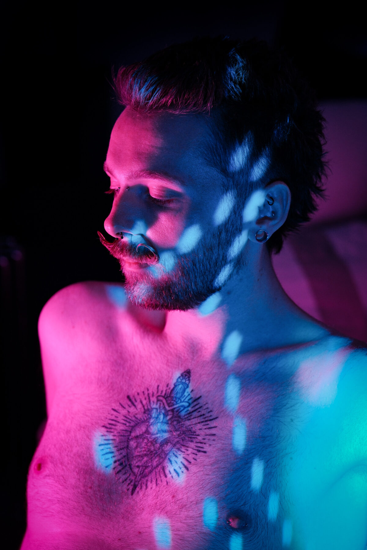 Portrait de François torse nu, jeux de lumière colorée, moustache en guidon, tatouage sur le torse, plug à l'oreille