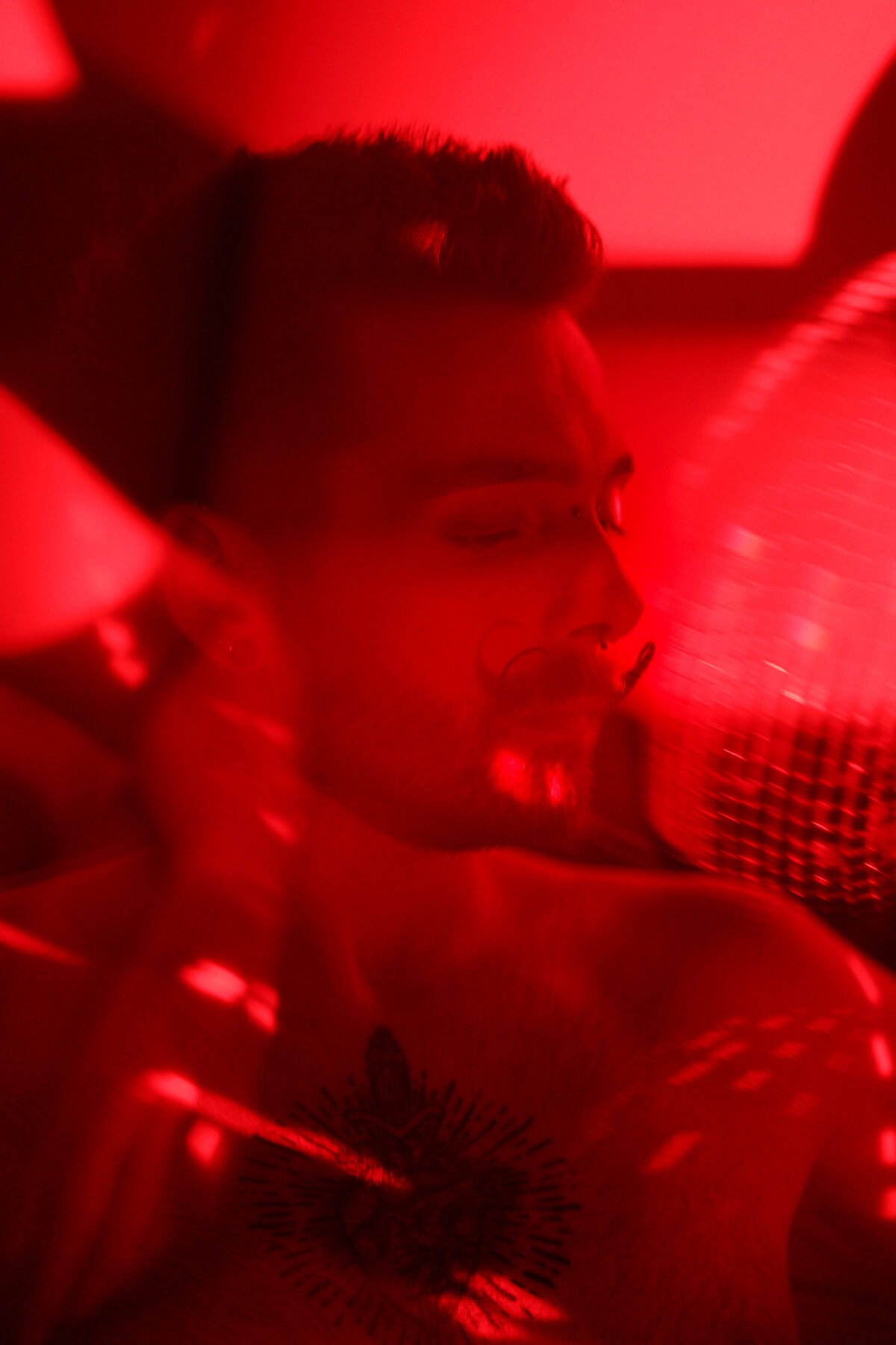Portrait de François sous lumière rouge, yeux fermés, boule à facettes floue en arrière-plan, torse nu
