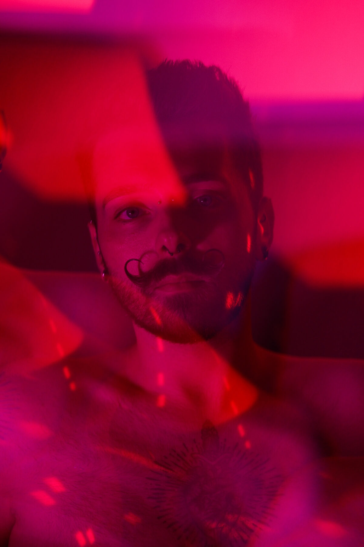 Portrait de François sous lumière rose et rouge, torse nu, tatouage sur le torse, moustache en guidon, piercings