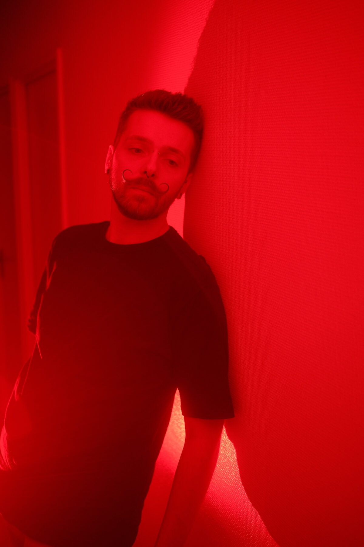 Portrait de François sous une lumière rouge, adossé à un mur, tee-shirt noir, regard vers l'objectif