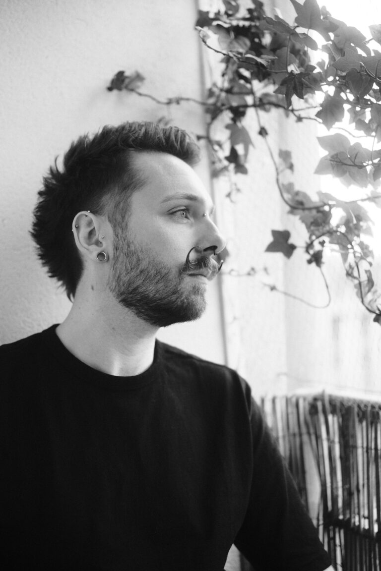 Portrait noir et blanc de François de profil contre un mur couvert de lierre, chemise noire, piercings à l'oreille