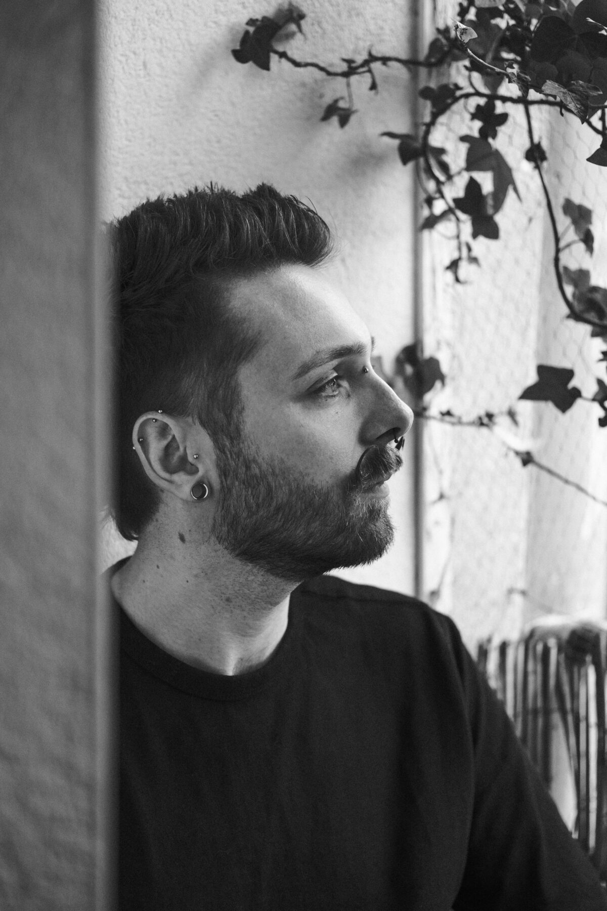 Portrait noir et blanc de François appuyé contre un mur couvert de lierre, chemise noire, plug à l'oreille