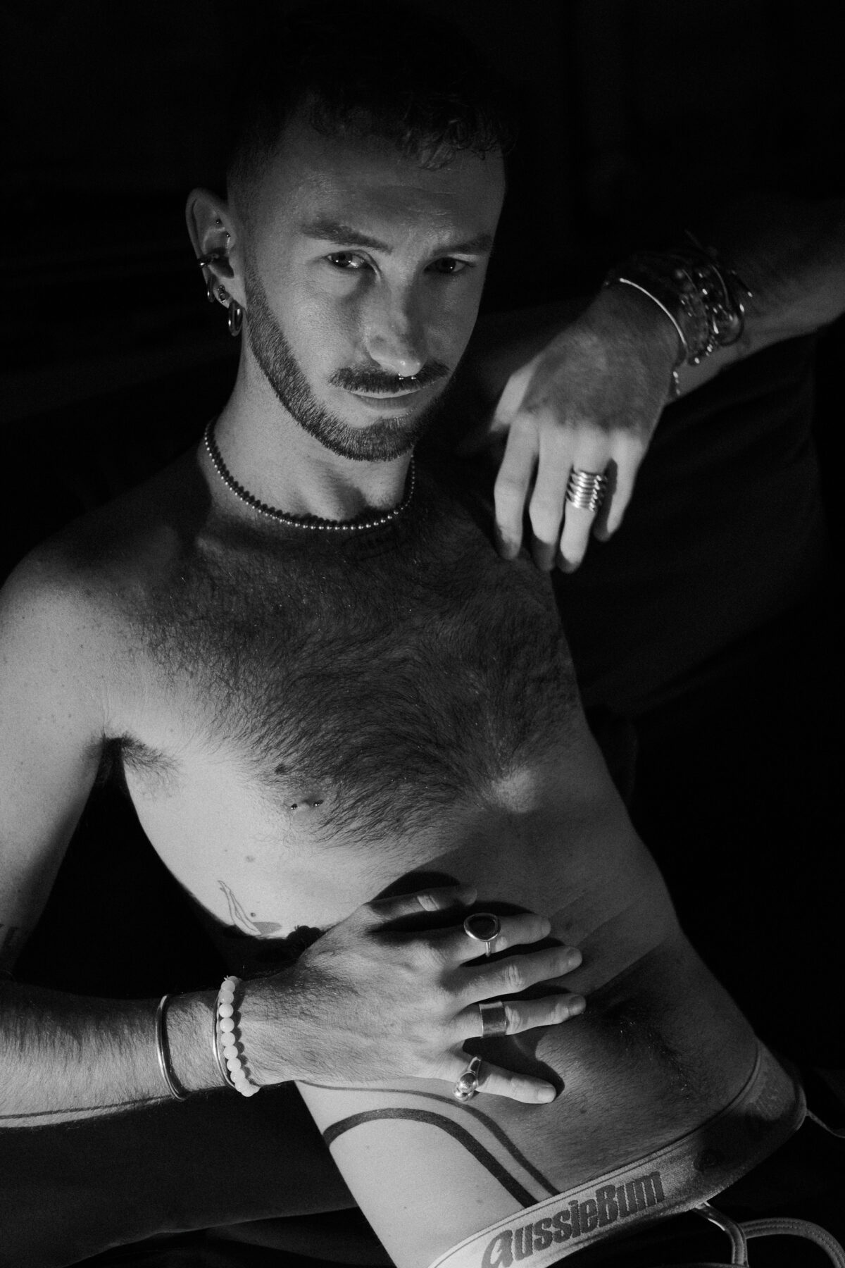 Gros plan en noir et blanc sur le torse de Hugo (de Lille), mains ornées de bagues, sous-vêtement, tatouage
