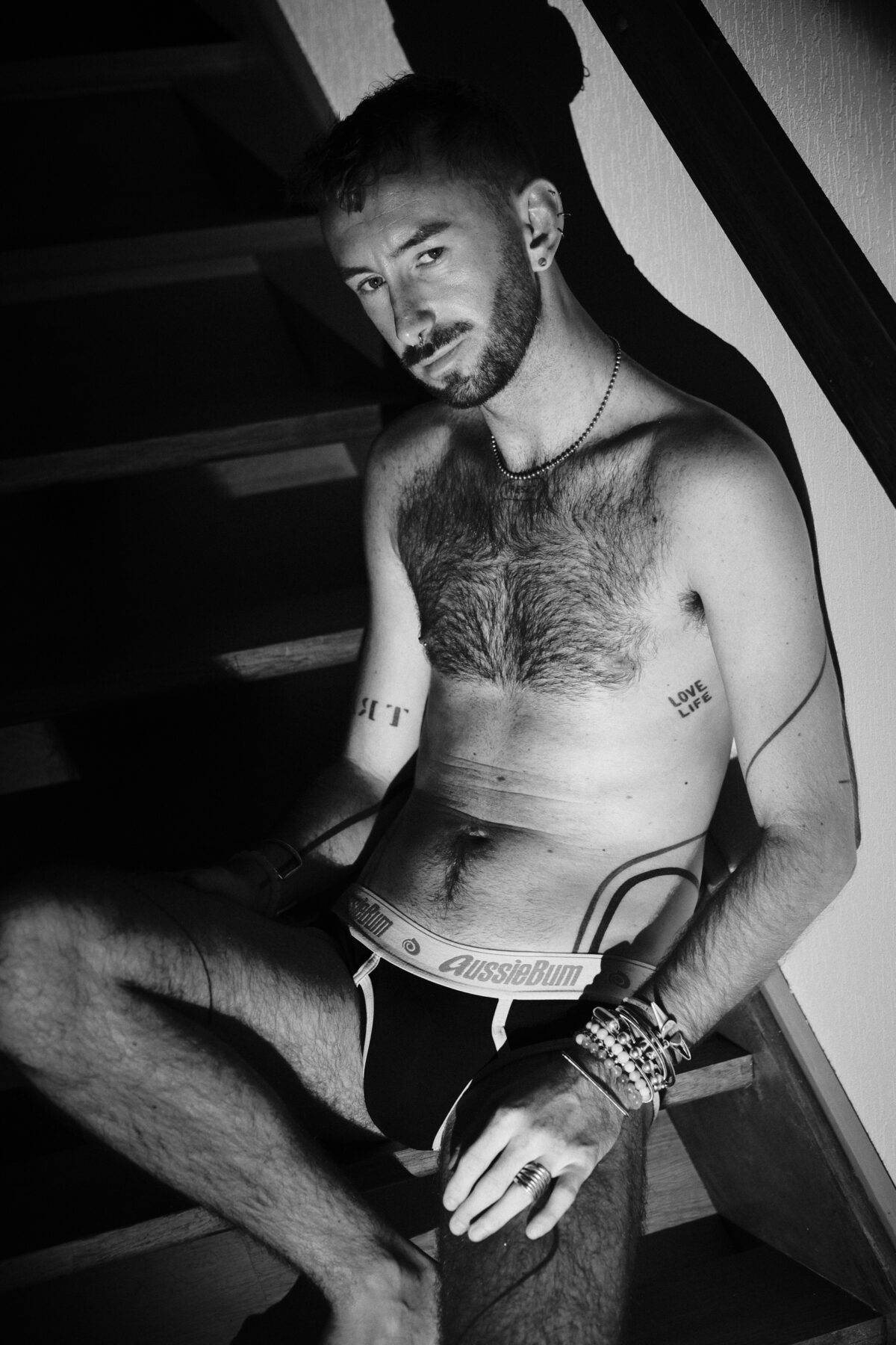 Portrait de Hugo (de Lille) assis dans un escalier, torse tatoué, sous-vêtement, bijoux et piercings, noir et blanc