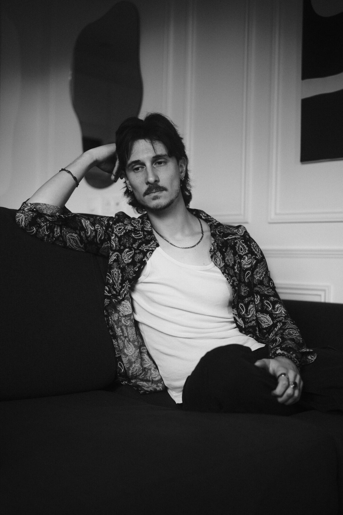 Portrait noir et blanc de Nico assis sur un canapé, chemise paisley ouverte sur débardeur blanc, bras levé derrière la tête
