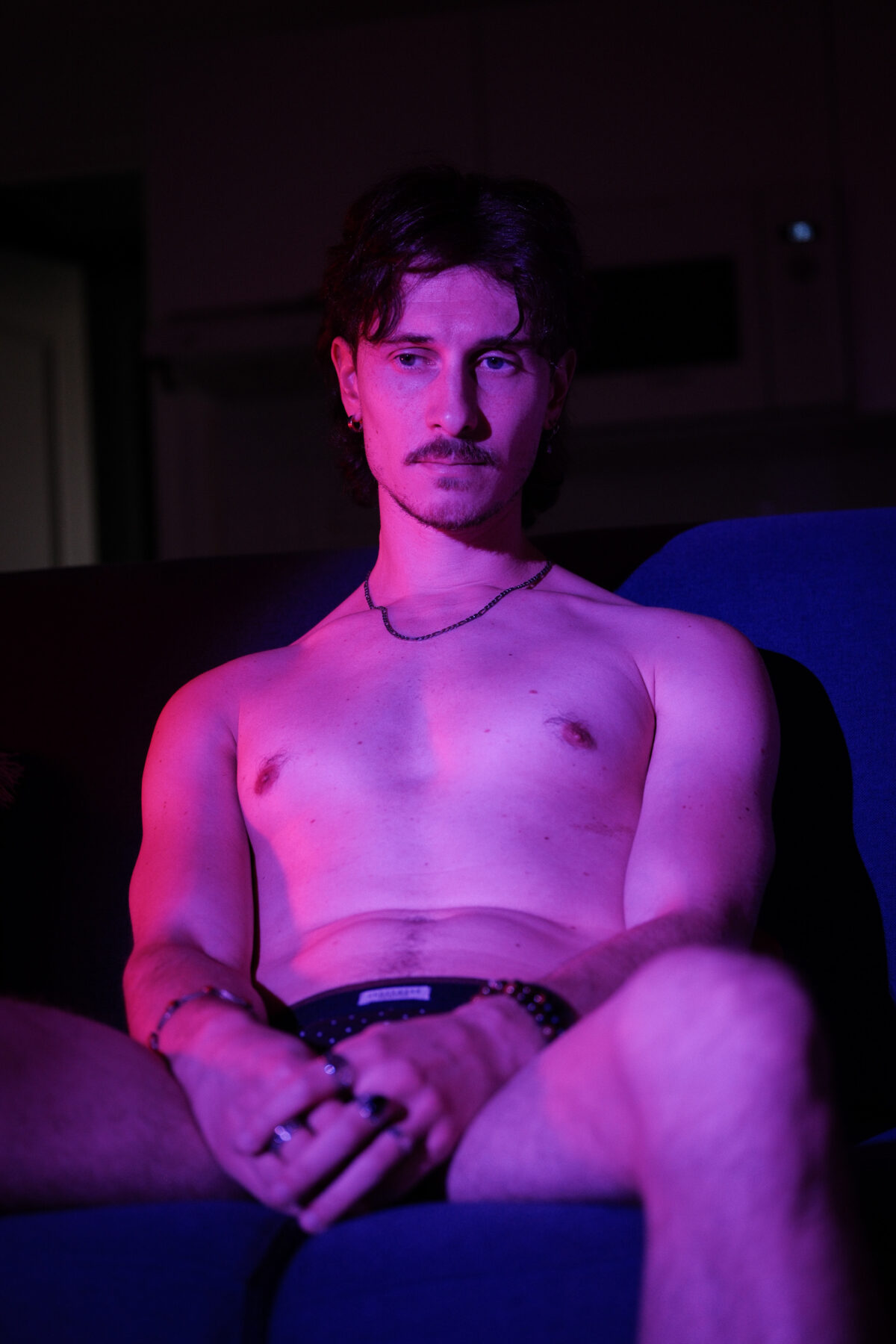 Portrait de Nico assis en sous-vêtement noir à pois, torse nu, éclairage rose et violet, collier visible